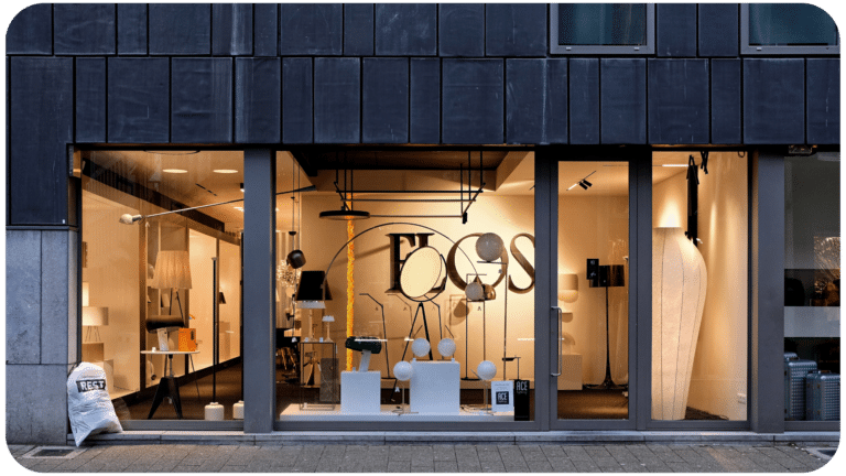 A modern store facade lit up at dusk, showcasing various designer lamps and furniture through a large glass window.